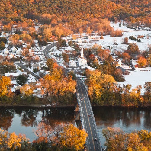 A scenic view of a snowy village with a river, trees, and a church, surrounded by autumn foliage at sunset.