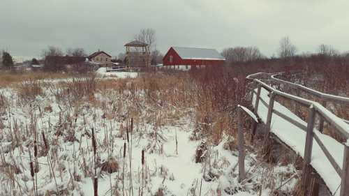 A snowy landscape with a wooden walkway, red barn, and distant buildings under a cloudy sky.