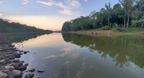 A serene river scene at sunset, with trees lining the banks and a person fishing on the shore.
