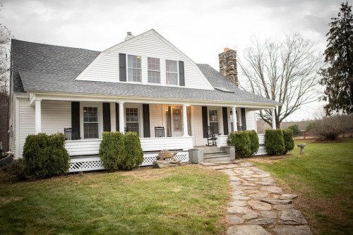 A charming white house with a stone pathway, surrounded by greenery and trees, under a cloudy sky.