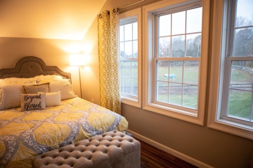 Cozy bedroom with a patterned yellow bedspread, a decorative pillow, and large windows overlooking a grassy field.