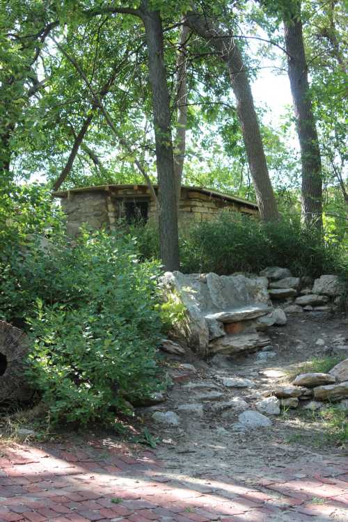 A stone structure nestled among trees, with a rocky path and a bench made of stone in a natural setting.