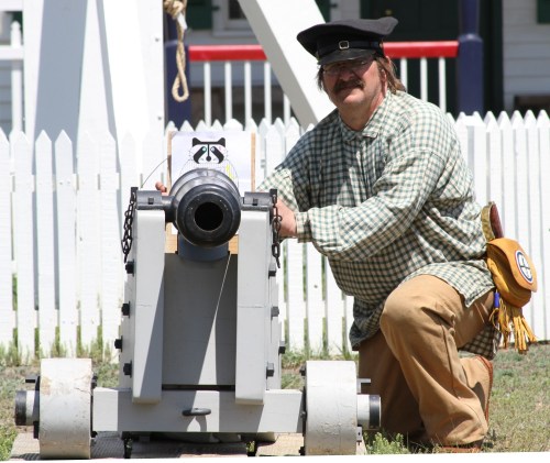 A person in historical attire kneels beside a cannon, preparing it for use, with a white picket fence in the background.