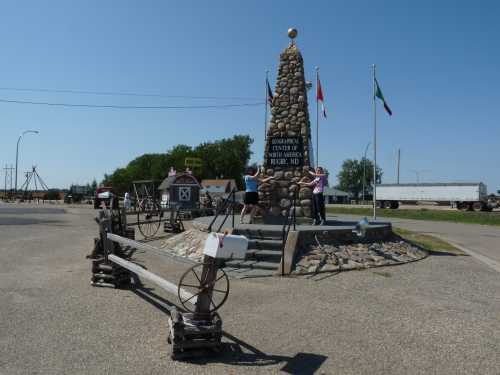 A stone monument with flags, surrounded by historical artifacts, and two people posing for a photo in a sunny area.