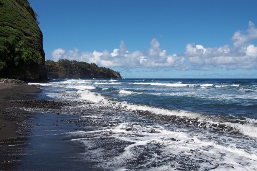A scenic beach with black sand, gentle waves, and lush green cliffs under a bright blue sky with scattered clouds.