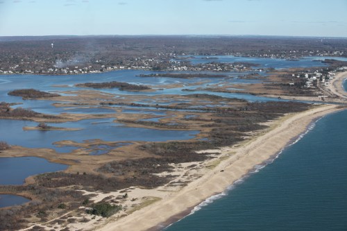 Aerial view of a coastal landscape featuring sandy beaches, wetlands, and calm blue waters under a clear sky.