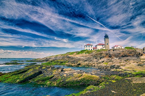 A scenic view of a lighthouse on rocky shores under a vibrant blue sky with wispy clouds.