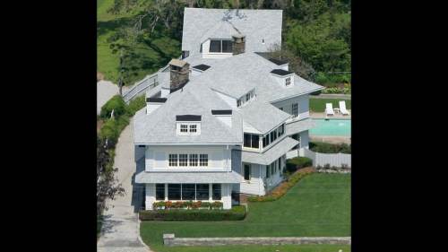 Aerial view of a large, multi-story house with a gray roof, surrounded by green lawn and a swimming pool.