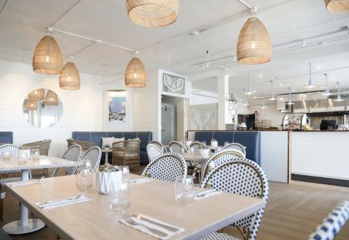 Bright, modern restaurant interior with woven pendant lights, wooden tables, and blue accents. Cozy seating area visible.