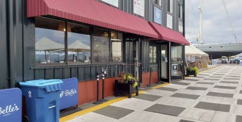 Exterior view of a café with a red awning, outdoor seating, and a blue trash bin near a waterfront.
