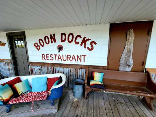 Sign for "Boon Docks Restaurant" with colorful cushions on a vintage couch and wooden bench in a rustic setting.