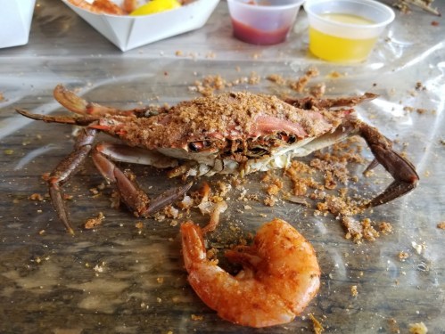 A seasoned crab with breadcrumbs sits on a table next to a cooked shrimp and small cups of sauce.