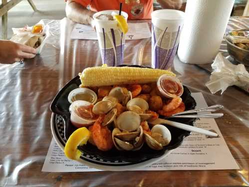 A seafood platter with shrimp, clams, corn on the cob, and dipping sauces, served on a black plate.