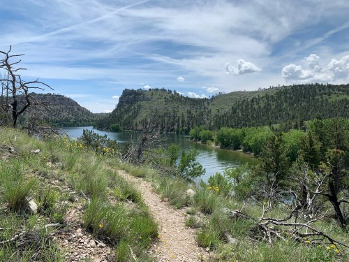 A scenic river view surrounded by green hills and trees under a partly cloudy sky, with a winding path in the foreground.