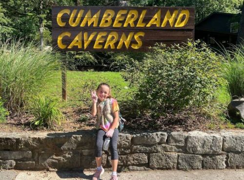 A child smiles and waves in front of a sign that reads "Cumberland Caverns," surrounded by greenery.