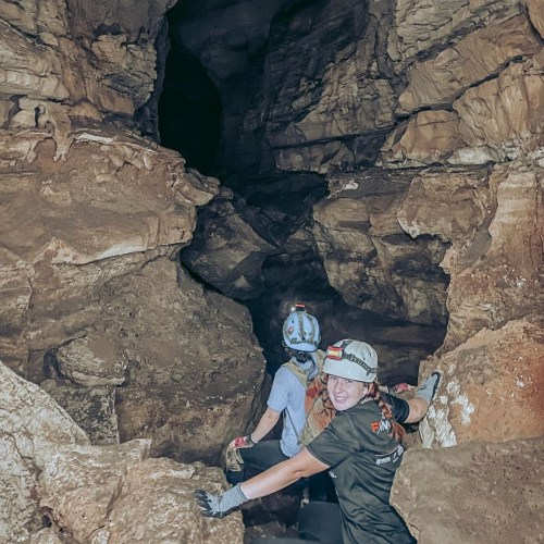 Two people in helmets exploring a cave, surrounded by rocky walls and dark passages.