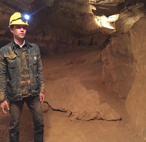 A person wearing a hard hat with a headlamp stands in a dimly lit cave, surrounded by rocky terrain.