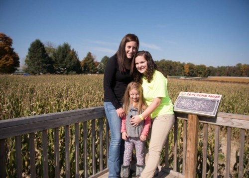 A woman and two girls smile together on a platform overlooking a corn maze on a sunny day.