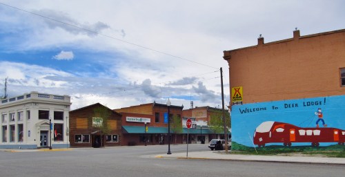 A small town street scene featuring a mural welcoming visitors to Deer Lodge and historic buildings under a cloudy sky.