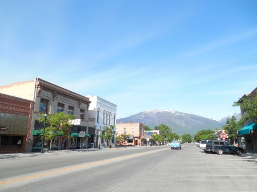 A quiet street scene with historic buildings, trees, and mountains in the background under a clear blue sky.