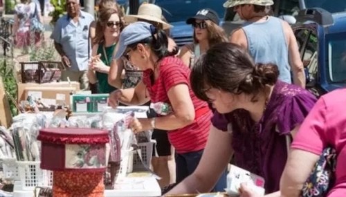 A busy outdoor market scene with people browsing tables filled with various items on a sunny day.