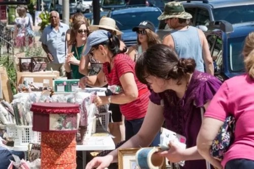 A bustling outdoor market scene with people browsing tables filled with various items and artwork.