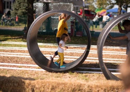 A child runs inside a large metal wheel while another child plays nearby in a sunny outdoor setting.