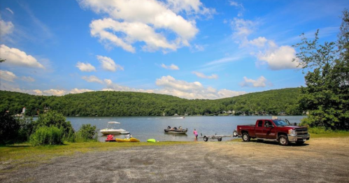 A serene lake scene with boats, a red truck parked nearby, and lush green hills under a blue sky with fluffy clouds.