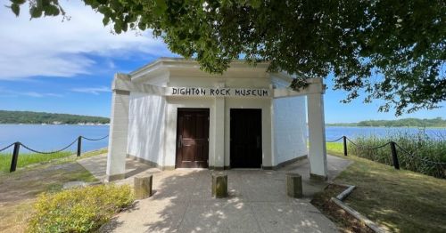Exterior view of the Dighton Rock Museum, featuring a white building by a lake, surrounded by greenery and a clear blue sky.