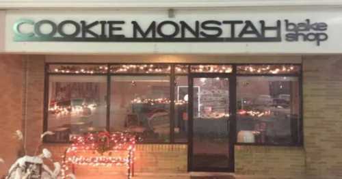 Sign for "Cookie Monstah Bake Shop" with festive lights, showcasing a cozy storefront in a snowy setting.