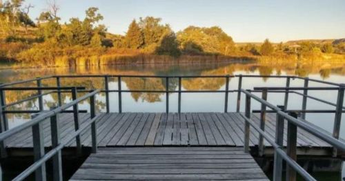 A wooden dock extends over a calm lake, surrounded by trees and reflecting the serene landscape.