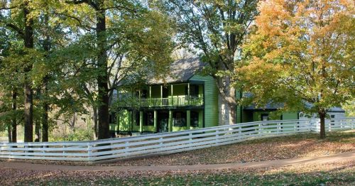 A green house surrounded by trees with autumn foliage and a white fence, set in a serene landscape.