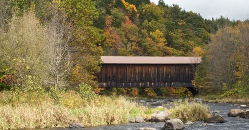 A rustic covered bridge spans a river, surrounded by vibrant autumn foliage and rocky riverbanks.