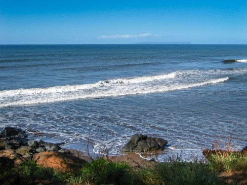 A serene beach scene with gentle waves, rocky shore, and a clear blue sky in the background.