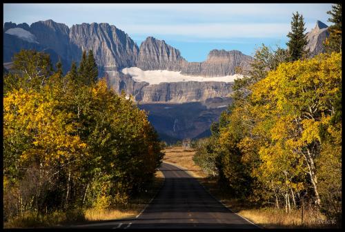 A scenic road lined with autumn trees leads to majestic mountains under a clear blue sky.