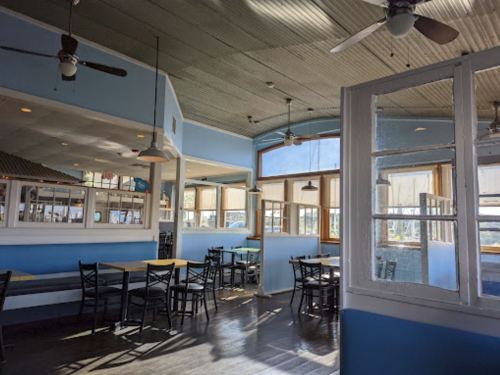 Bright, airy restaurant interior with blue walls, wooden tables, and ceiling fans, featuring large windows for natural light.