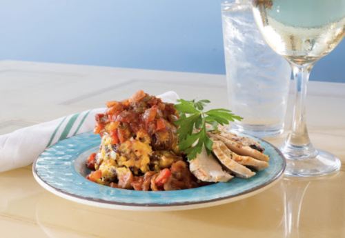 A colorful plate of layered casserole topped with salsa, accompanied by sliced chicken and a glass of water.