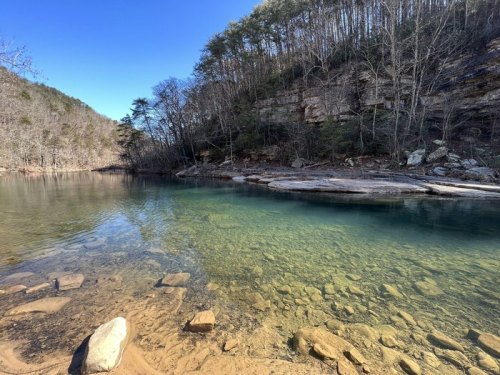 A serene river scene with clear water, rocky shore, and trees lining the banks under a blue sky.
