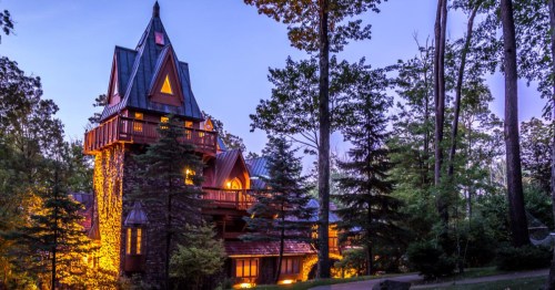 A tall, whimsical house with a pointed roof, surrounded by trees, illuminated at dusk.
