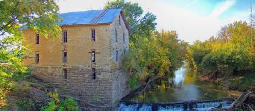 Historic stone building beside a calm river, surrounded by lush greenery and trees under a clear blue sky.