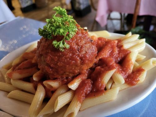 A plate of penne pasta topped with marinara sauce and a large meatball, garnished with parsley.