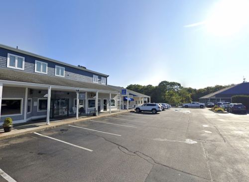A sunny day at a shopping plaza with parked cars and a two-story building in the background.