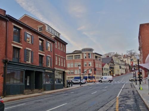 A street view featuring brick buildings, a sloped road, and a mix of modern and historic architecture under a cloudy sky.