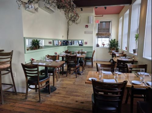 Cozy restaurant interior with wooden tables, chairs, and plants, featuring large windows and a warm atmosphere.