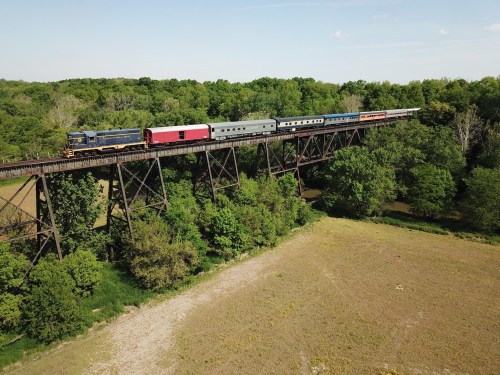 A train crosses a tall trestle bridge surrounded by lush greenery and open fields under a clear blue sky.
