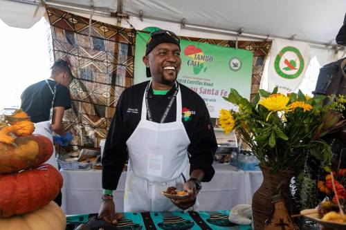 A smiling chef in a black uniform serves food at a vibrant festival booth decorated with flowers and pumpkins.