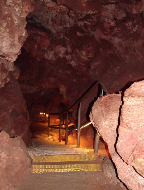 A dimly lit cave interior with a metal staircase leading down, surrounded by rocky walls.
