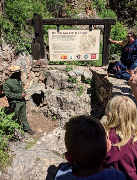 A park ranger stands by a historical sign, with visitors seated nearby, in a natural setting with rocky terrain.