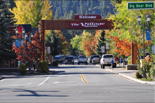 A welcoming archway reads "The Village" in a colorful autumn setting, with cars parked along a tree-lined street.