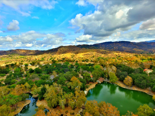 Aerial view of a serene landscape with a green lake, lush trees, and rolling hills under a partly cloudy sky.